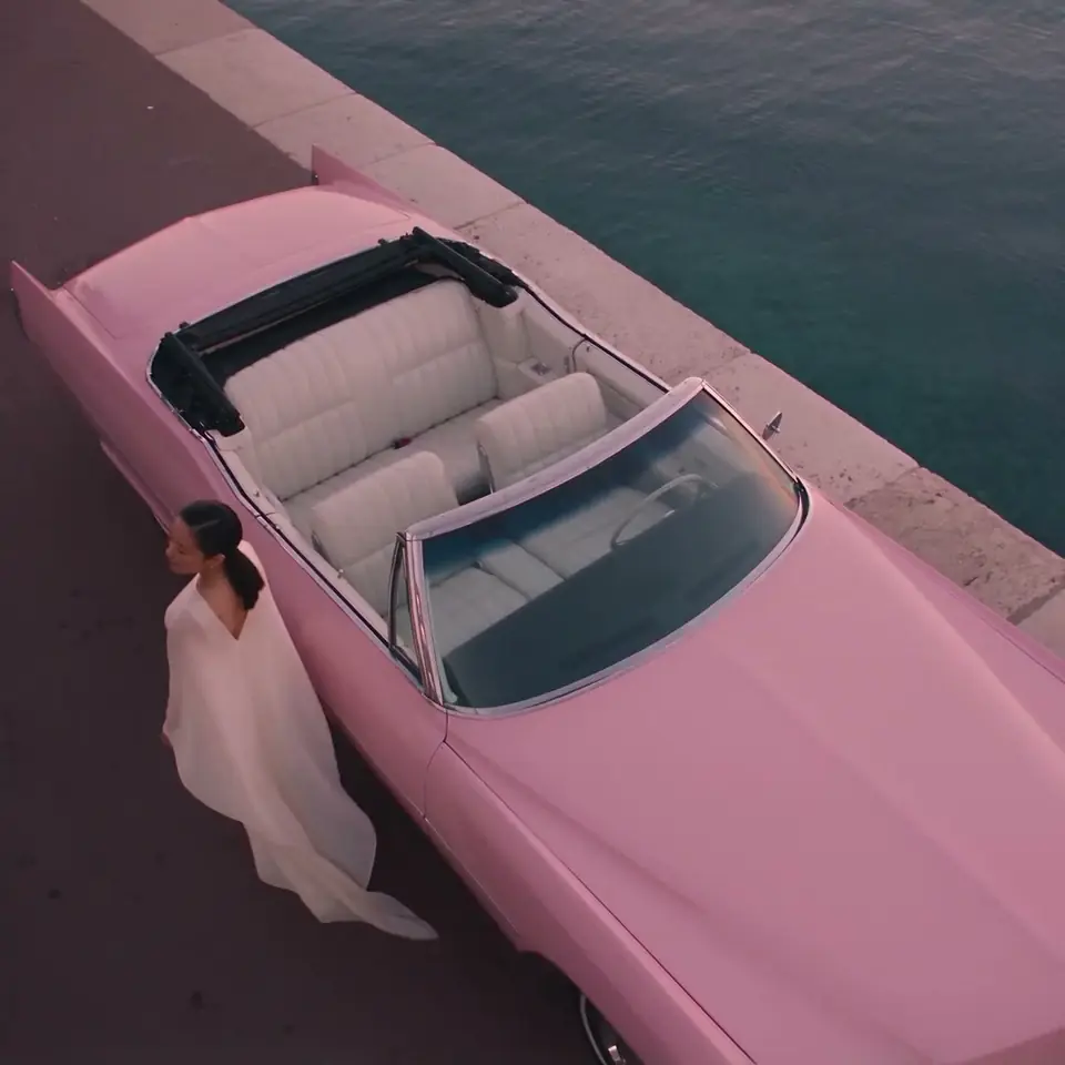 A pink convertible parked by the sea, seen from above.